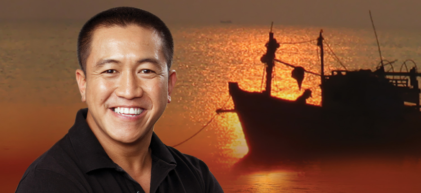 Comedian and author Anh Do is pictured smiling at the camera face on. In the background is a sea at sunrise with a boat floating calmly on the still ocean.
